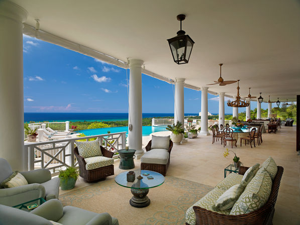 Oceanfront terrace with white columns, wicker lounge seating, infinity pool and panoramic turquoise sea view, plus al fresco dining under chandeliers and ceiling fans
