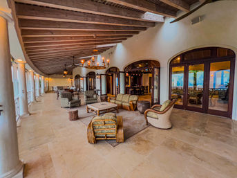 Ocean-view Mediterranean-style covered terrace with exposed wooden beams, columned walkway, rattan and upholstered lounge seating, chandelier lighting and arched glass doors to an indoor bar.