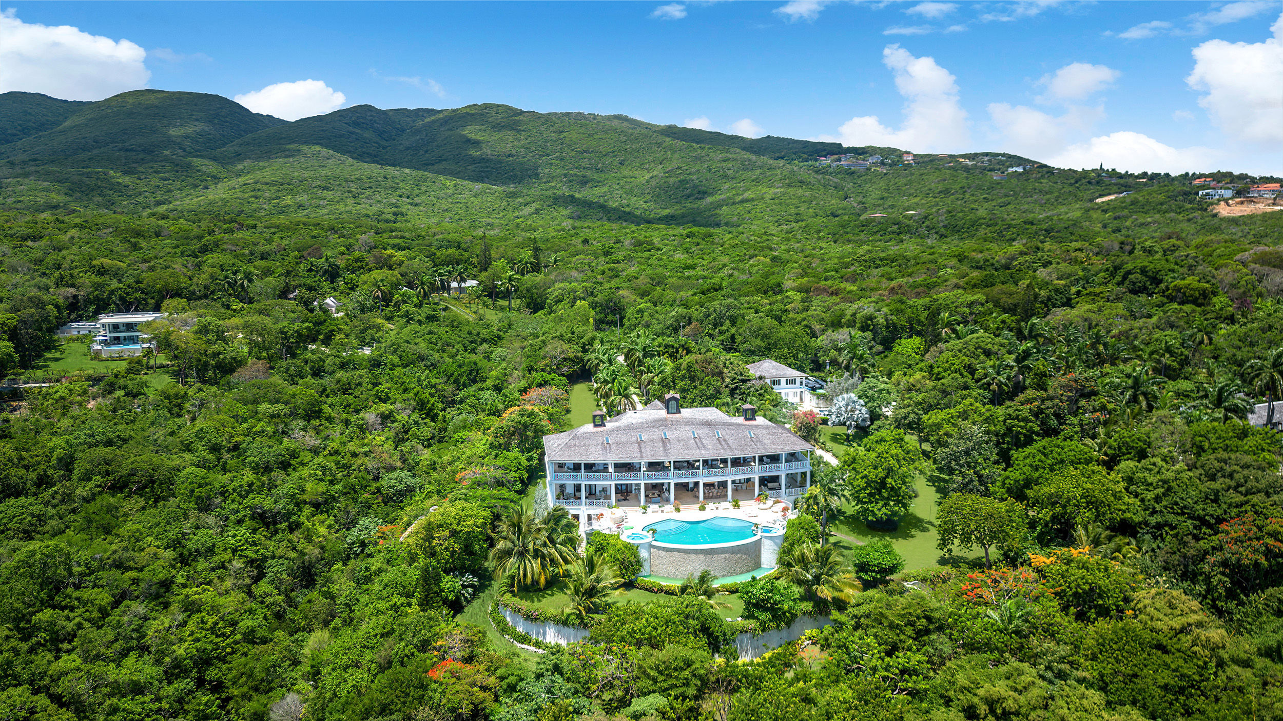 Aerial view of a luxury tropical hillside villa nestled in lush green forest, featuring a round infinity pool and sun-soaked terrace with rolling mountains and a bright blue sky.