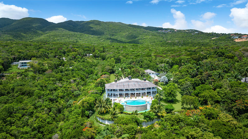 Aerial view of a luxury tropical hillside villa nestled in lush green forest, featuring a round infinity pool and sun-soaked terrace with rolling mountains and a bright blue sky.