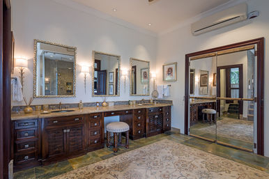 Spacious spa-like master bathroom with dark wood double vanity and marble countertop, three ornate framed mirrors, wall sconces, upholstered vanity stool, patterned area rug, and mirrored sliding doors reflecting a walk-in glass shower.