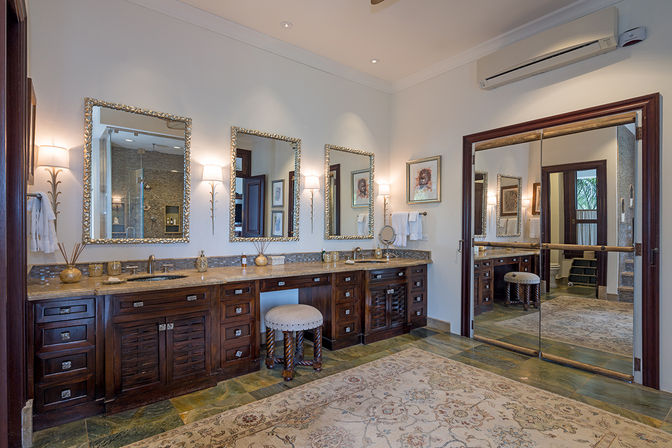 Spacious spa-like master bathroom with dark wood double vanity and marble countertop, three ornate framed mirrors, wall sconces, upholstered vanity stool, patterned area rug, and mirrored sliding doors reflecting a walk-in glass shower.