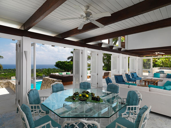Bright open-plan tropical villa with dark wooden ceiling beams and fan, glass-top dining table surrounded by teal-cushioned wicker chairs, airy white sofas and blue accent chairs, folding doors opening to a stone-walled pool terrace, palm trees and distant ocean view.