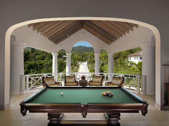 Billiards table on a sunlit villa veranda with cushioned chairs and white arched columns, overlooking a palm-lined stone pathway through lush tropical gardens toward green hills.