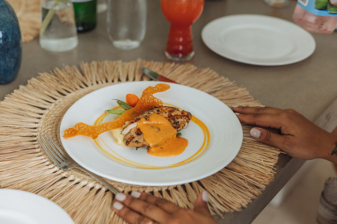 Grilled herb chicken breast drizzled with bright orange sauce on mashed potatoes, topped with a crisp cheese tuile and green beans on a white plate set on a woven straw placemat, hands nearby and a red cocktail in the background.