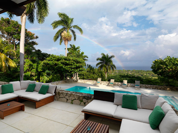 Sunlit tropical patio with white-cushioned wooden lounge and green pillows beside a stone-edged swimming pool, palm trees framing a rainbow arching over the distant ocean — a relaxing vacation-ready view.