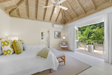 Bright airy bedroom with vaulted wood ceiling and ceiling fan, king bed dressed in white with green accent pillows, rattan chair and bench, sliding glass doors opening to a sunlit balcony overlooking lush tropical greenery — a serene coastal retreat.