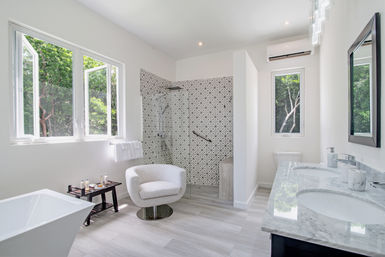 Bright spa-like modern bathroom with marble double vanity, freestanding tub, patterned tile walk-in shower, cozy swivel chair and large windows framing lush green trees.