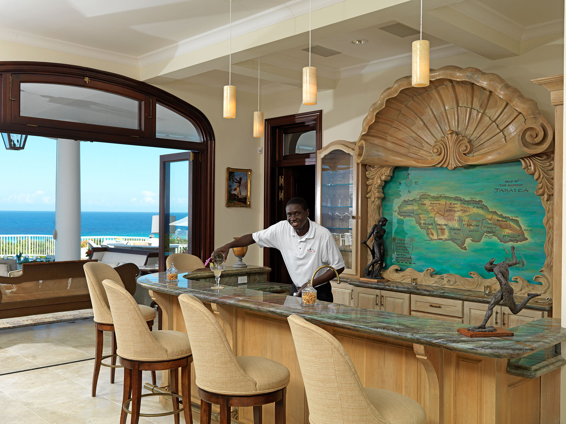 Ocean-view indoor bar in a tropical villa with marble countertop and pendant lights; a bartender pours a cocktail at a curved wooden bar framed by a large carved shell and colorful map of Jamaica, cushioned barstools and blue sea visible through arched doors.
