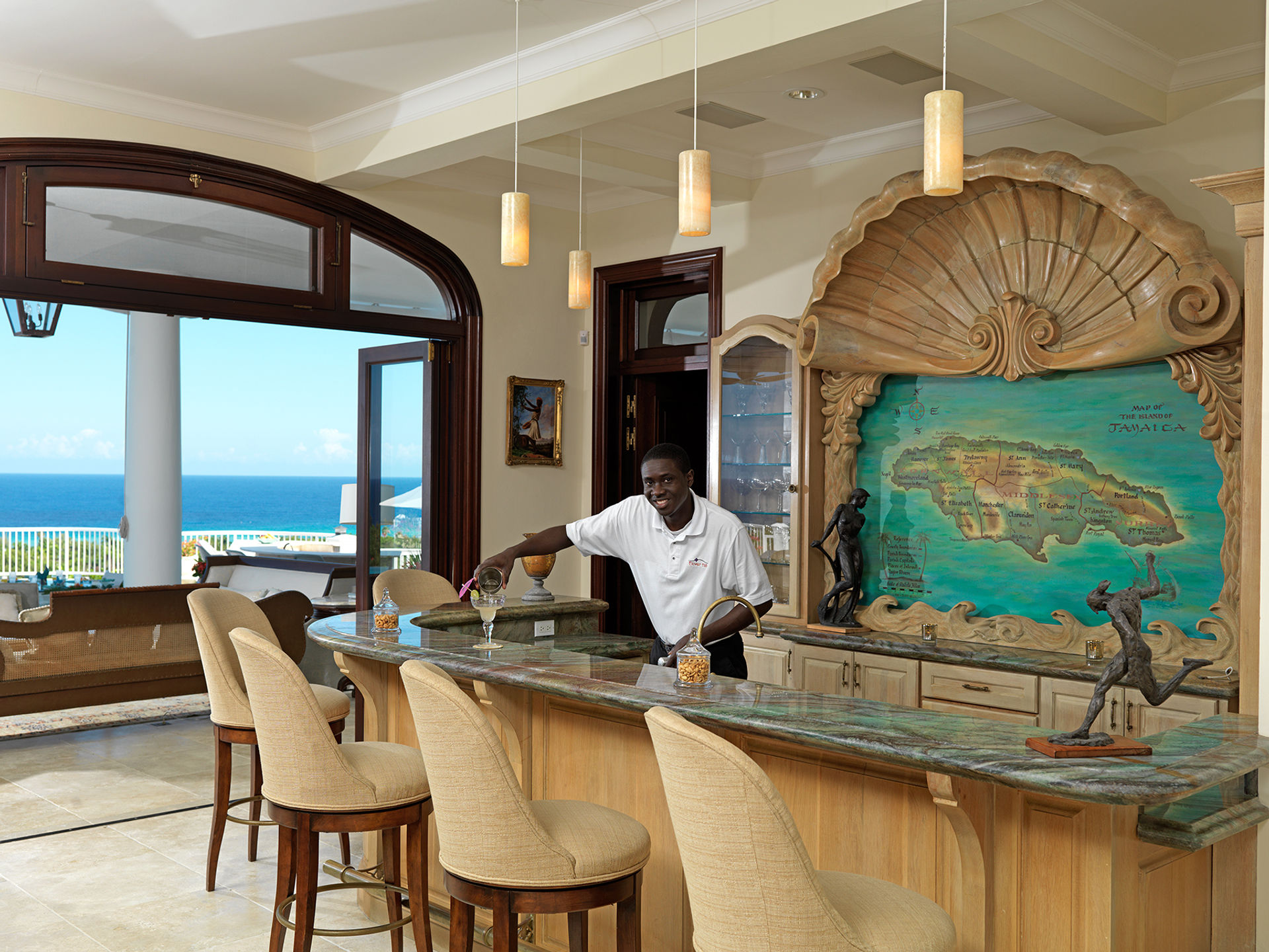 Ocean-view indoor bar in a tropical villa with marble countertop and pendant lights; a bartender pours a cocktail at a curved wooden bar framed by a large carved shell and colorful map of Jamaica, cushioned barstools and blue sea visible through arched doors.