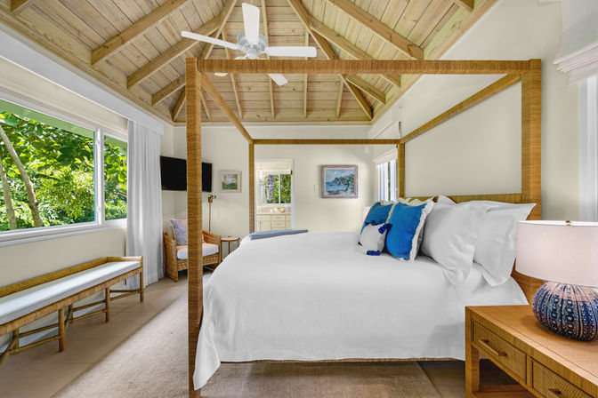 Airy sunlit tropical bedroom with rattan four‑poster canopy bed in white linens and blue accent pillows, vaulted wood ceiling with fan, wicker seating and window bench overlooking lush greenery, bedside lamp and visible en‑suite — coastal vacation rental vibe.