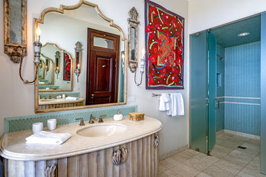 Bright luxury bathroom with a curved marble vanity and oval sink, ornate scalloped mirror, teal mosaic backsplash, glass-door turquoise tile shower, colorful red wall textile and white towels.