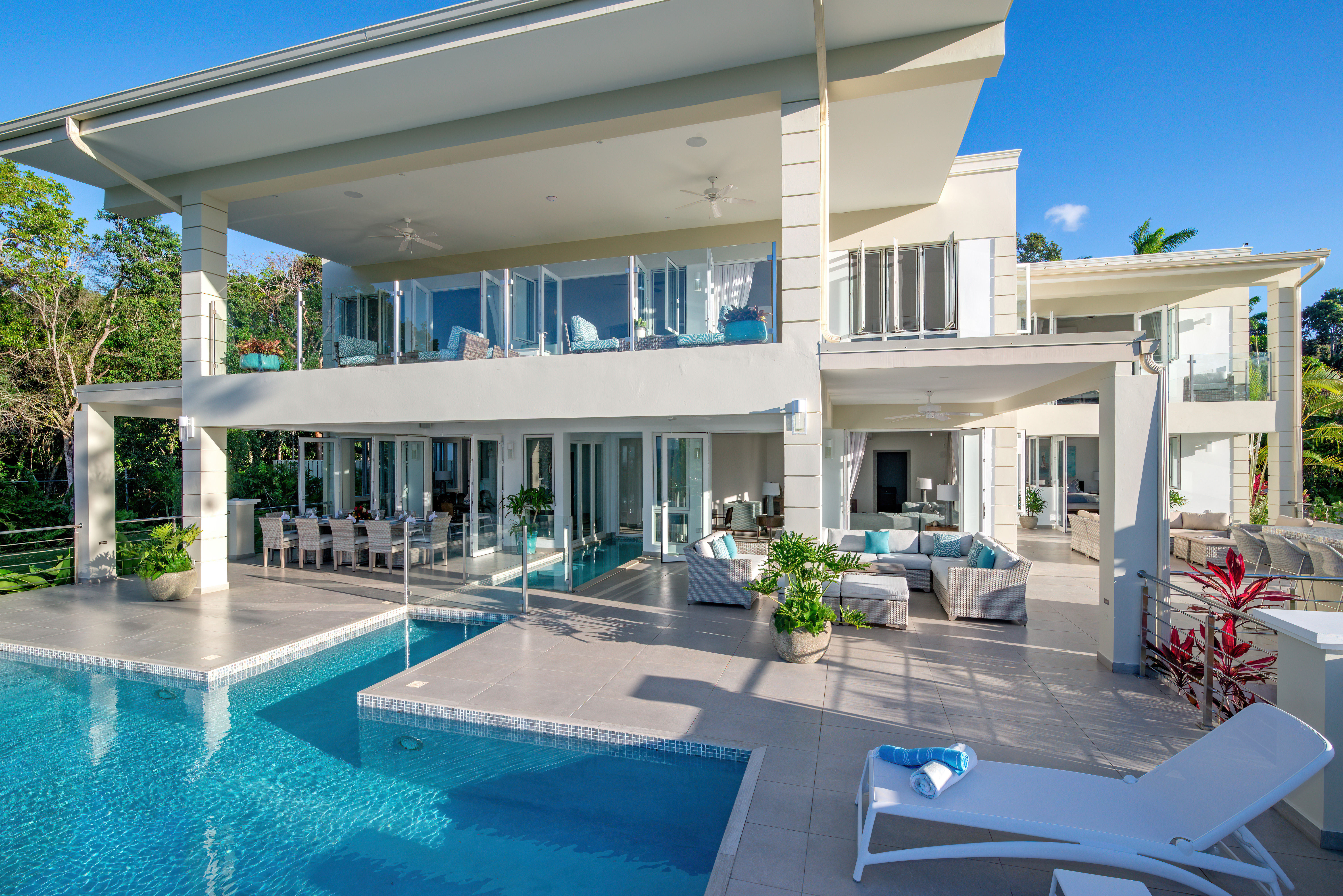 Sunlit modern two-story luxury villa in a tropical setting with a turquoise pool, tiled poolside patio and loungers, outdoor dining and wicker lounge seating, glass balcony and lush greenery
