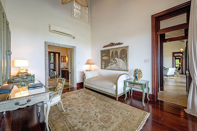 Airy sunlit villa bedroom sitting area with hardwood floors, tufted chaise lounge, vintage mirrored desk, ornate framed artwork and green side table with peacock sculpture