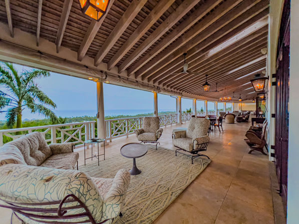 Relaxing ocean-view tropical veranda — spacious covered outdoor lounge with cushioned sofas and armchairs, ceiling fans and lanterns, tiled floor and palm trees visible beyond the white railing