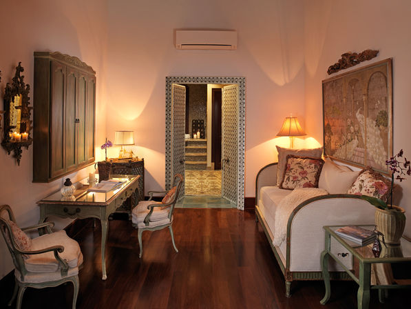 Cozy Mediterranean-inspired sitting room with carved wood furniture, cream sofa with floral cushions, glass-top desk, warm lamp lighting and dark hardwood floors, patterned doors opening to a tiled bath.