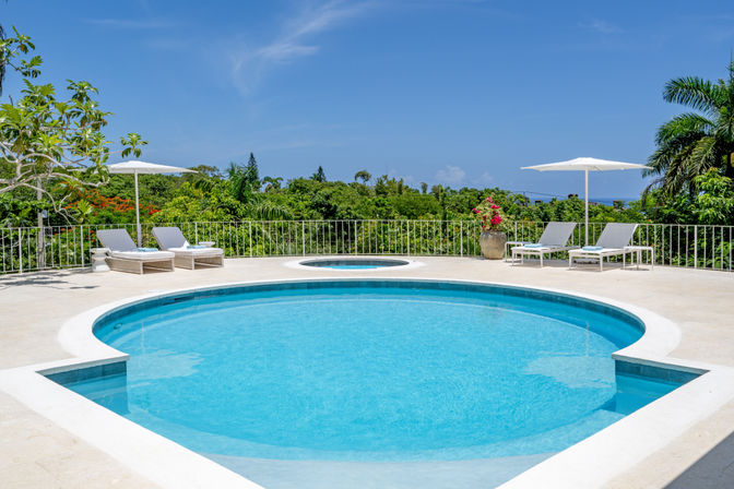 Sun-drenched circular swimming pool and spa on a tropical terrace with white loungers, umbrellas, palm trees and a distant ocean view.
