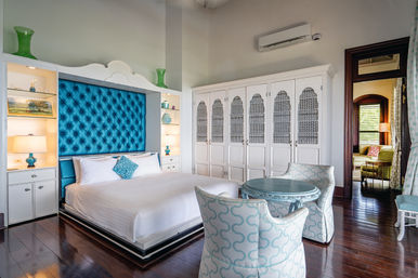 Coastal-chic boutique hotel bedroom with tufted turquoise headboard, crisp white bedding, built-in shelves and lamps, large white armoire, round turquoise table and patterned chairs on dark hardwood floors.