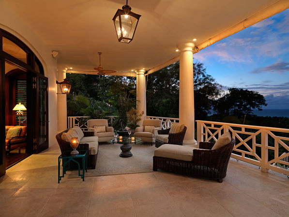 Ocean-view tropical veranda at dusk with columned porch, wicker lounge chairs and cushioned seating around a glass coffee table, warm lantern lighting and ceiling fan overlooking trees and the sea.