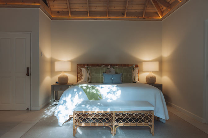 Cozy neutral bedroom with vaulted wood ceiling, king bed with green pillows, rattan bench at foot, matching bedside lamps and dappled sunlight on white bedding.