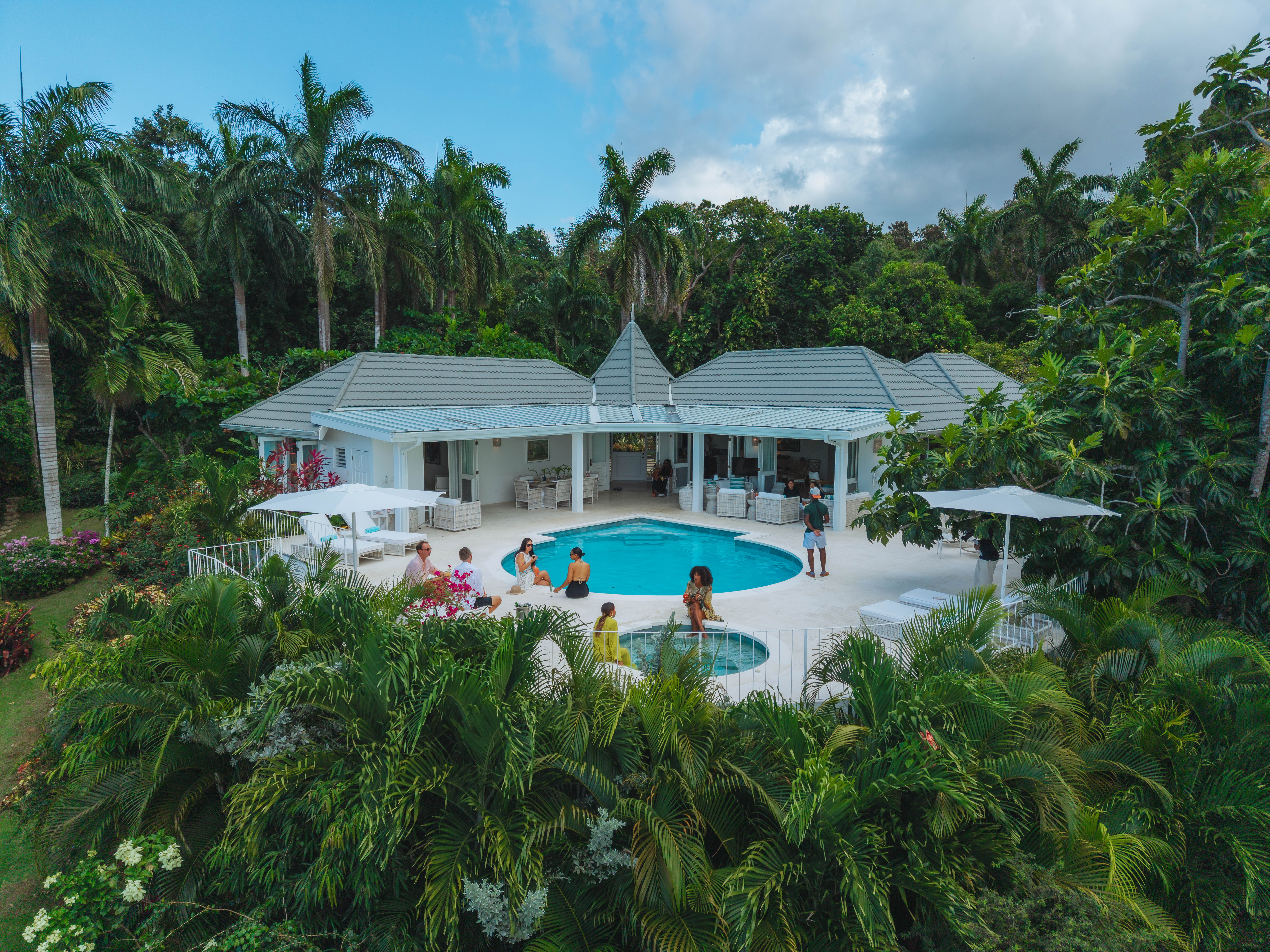 Aerial view of a white luxury tropical villa with a curved swimming pool and hot tub, surrounded by lush palm trees and guests relaxing on the patio.