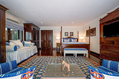 Spacious luxury bedroom interior with dark wood four‑poster king bed, navy blue and white coastal textiles, daybed and matching armchairs, patterned blue area rug, hardwood floors, wooden armoire and TV cabinet, and a glass coffee table topped by a small dog sculpture