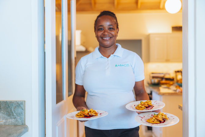 Smiling server in a white polo holding three plates of golden shrimp appetizers with sauce, standing in a bright modern kitchen doorway.