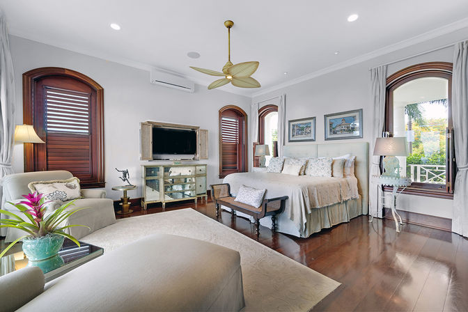 Bright coastal-style master bedroom with king bed, hardwood floors, arched wooden windows with shutters, cozy seating area, ceiling fan, mirrored TV console, bedside lamps, and a potted tropical plant flooding the room with natural light.
