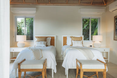 Sunlit tropical twin bedroom with white linens and fringed throws, rattan headboards, matching white bedside tables and round textured lamps, wooden benches at the foot of each bed, lush greenery visible through two windows.