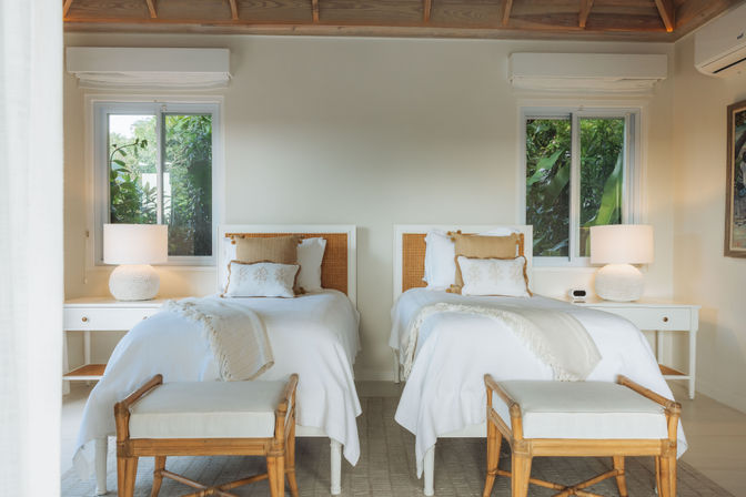 Sunlit tropical twin bedroom with white linens and fringed throws, rattan headboards, matching white bedside tables and round textured lamps, wooden benches at the foot of each bed, lush greenery visible through two windows.