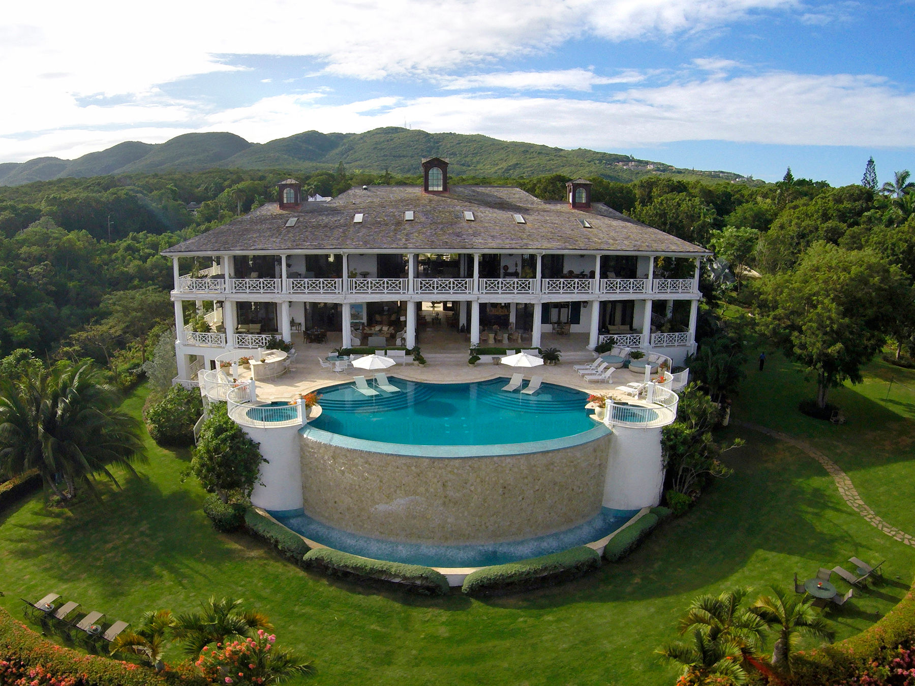 Aerial view of a luxury three-level villa on a tropical hillside with wraparound verandas and a curved infinity pool overlooking lush green hills and distant mountains.