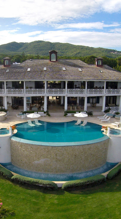 Aerial view of a luxury three-level villa on a tropical hillside with wraparound verandas and a curved infinity pool overlooking lush green hills and distant mountains.