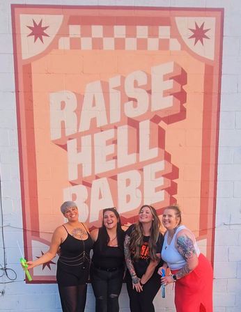 Four smiling friends strike a playful pose in front of a large pink retro brick wall mural reading "RAISE HELL BABE", wearing colorful outfits and showing tattoos in an urban outdoor setting.