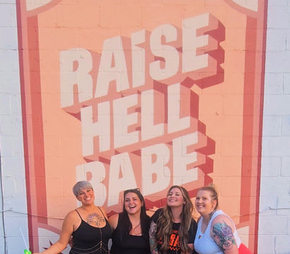 Four smiling friends strike a playful pose in front of a large pink retro brick wall mural reading "RAISE HELL BABE", wearing colorful outfits and showing tattoos in an urban outdoor setting.