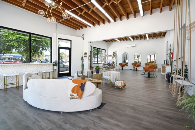 Bright, sunlit modern hair salon interior with exposed wooden beam ceiling, large windows, curved white lounge sofa, orange styling chairs, retail shelves and hanging swing seats.