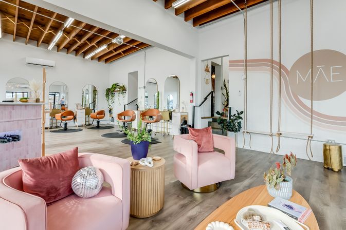 Bright modern boutique hair salon interior with exposed wood beams, blush-pink lounge chairs, brass-accented styling stations and round mirrors, lush potted plants, and playful rope swings along a white accent wall.