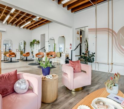 Bright modern boutique hair salon interior with exposed wood beams, blush-pink lounge chairs, brass-accented styling stations and round mirrors, lush potted plants, and playful rope swings along a white accent wall.