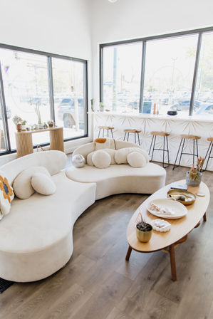Bright modern urban lounge with a curved cream sofa and round pillows, wooden oval coffee table with plants and décor, textured white bar with wooden stools and large street-facing windows — airy minimalist interior.