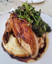 Roasted chicken breast with crispy skin atop creamy mashed potatoes, charred broccolini and microgreens drizzled with balsamic reduction on a white restaurant plate.