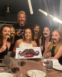 Group of adults at a home dinner party around a dining table, everyone smiling and holding novelty mustache props, woman in front holding a large chocolate mustache dessert with raspberries, wine glasses on table