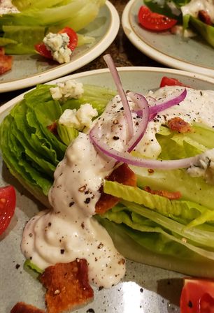 Classic wedge salad with a crisp iceberg lettuce wedge topped with creamy blue-cheese dressing, red onion rings, bacon crumbles, blue cheese crumbles and cherry tomato halves, finished with cracked black pepper on a plate.