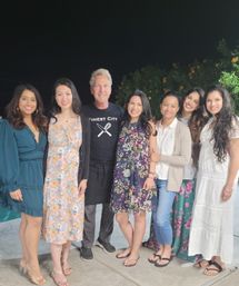 Seven people smiling in a nighttime backyard patio group photo, wearing casual summer dresses and sandals with a leafy garden backdrop