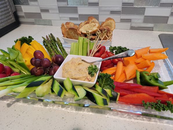 Vibrant veggie platter on a kitchen counter with creamy dip in the center surrounded by sliced cucumbers, carrots, red and green bell peppers, celery, asparagus, cherry tomatoes, grapes and crostini.
