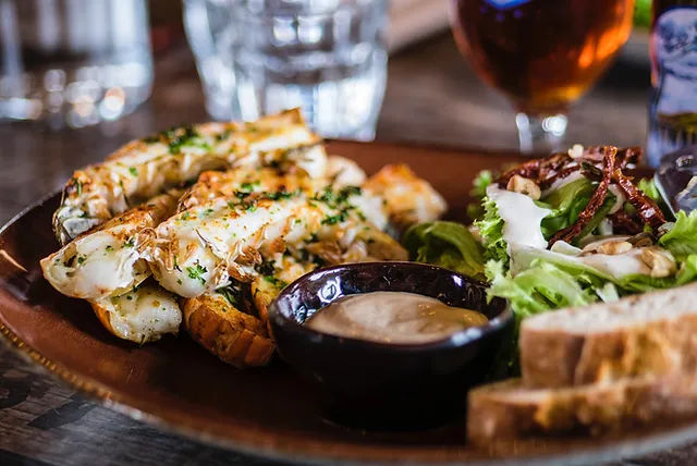 Close-up of grilled lobster tails sprinkled with herbs, a small bowl of creamy dipping sauce, mixed green salad and crusty bread on a wooden plate at a casual seafood restaurant.