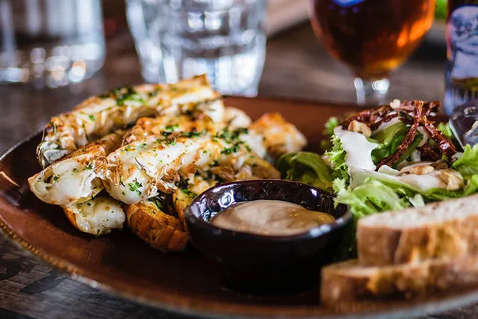 Close-up of grilled lobster tails sprinkled with herbs, a small bowl of creamy dipping sauce, mixed green salad and crusty bread on a wooden plate at a casual seafood restaurant.