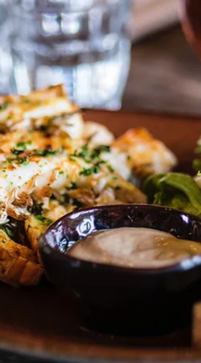Close-up of grilled lobster tails sprinkled with herbs, a small bowl of creamy dipping sauce, mixed green salad and crusty bread on a wooden plate at a casual seafood restaurant.