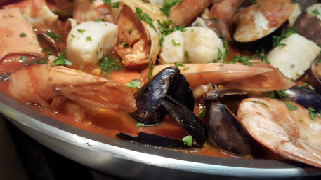 Close-up of Mediterranean-style seafood stew in a metal pan: whole shrimp, mussels, clams and scallops in a rich tomato broth, sprinkled with chopped parsley