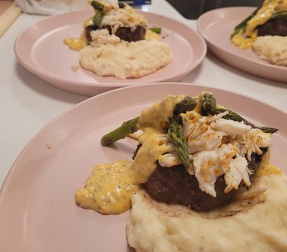 Gourmet beef patty topped with lump crab, asparagus and creamy hollandaise-style sauce served over mashed potatoes on a pink plate