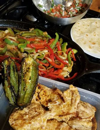 Colorful homemade fajita spread: cast-iron skillet of sizzling red and green bell peppers and onions, charred jalapeño halves, seasoned grilled chicken, flour tortillas and pico de gallo on a stovetop.