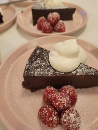 Close-up of a chocolate tart slice dusted with powdered sugar, topped with whipped cream and fresh raspberries on a pink dessert plate
